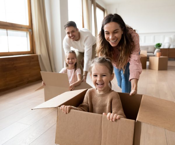 Happy parents playing with little kids riding in box Glückliche Familie die Umzugskarton durch die Wohnung schiebt.